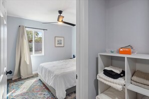 Una camera da letto con un letto, una ventola a soffitto, una finestra con tende, un quadro appeso al muro e uno scaffale con asciugamani e una scatola etichettata 'fogli essiccatori per bucato freschi'.