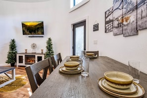 A dining area with a long table set for four, a fireplace, and a television mounted on the wall.