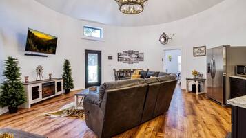 A modern living room with a large sectional sofa, a fireplace, a television mounted on the wall, and a kitchen area with a refrigerator and microwave.