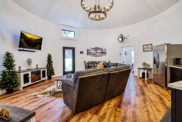 Una sala moderna con un sofá seccional grande, una chimenea, un televisor montado en la pared y una zona de cocina con un refrigerador y microondas.
