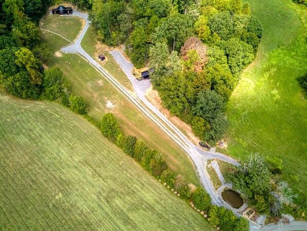 An aerial view of a rural property with a winding road, a small pond, and a cluster of buildings surrounded by dense greenery.
