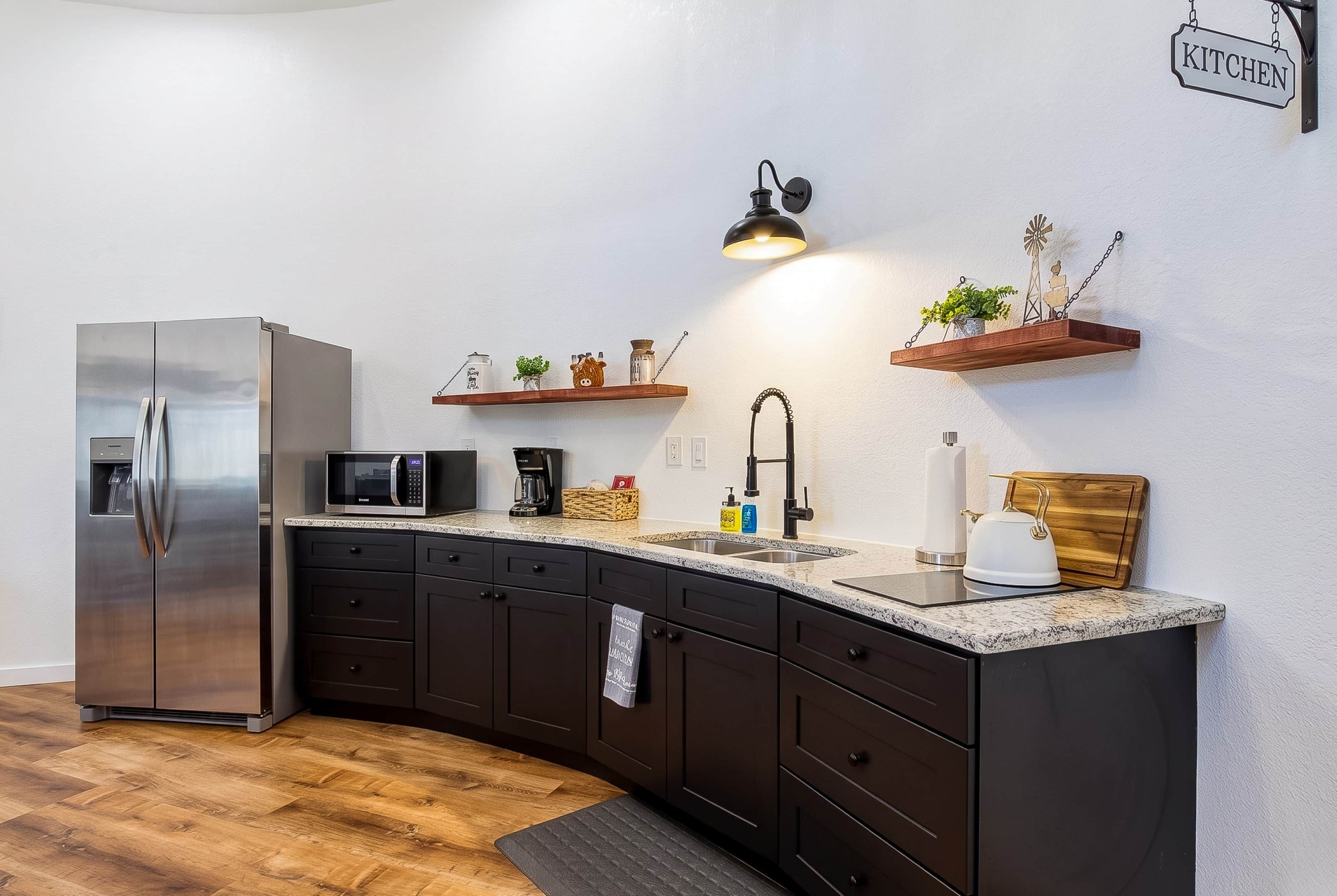 A modern kitchen with dark cabinetry, a stainless steel refrigerator, a microwave, and a sink with a black faucet. There are wooden shelves with decorative items and a hanging light fixture.