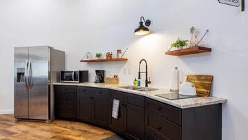 A modern kitchen with dark cabinetry, a stainless steel refrigerator, a microwave, and a sink with a black faucet. There are wooden shelves with decorative items and a hanging light fixture.