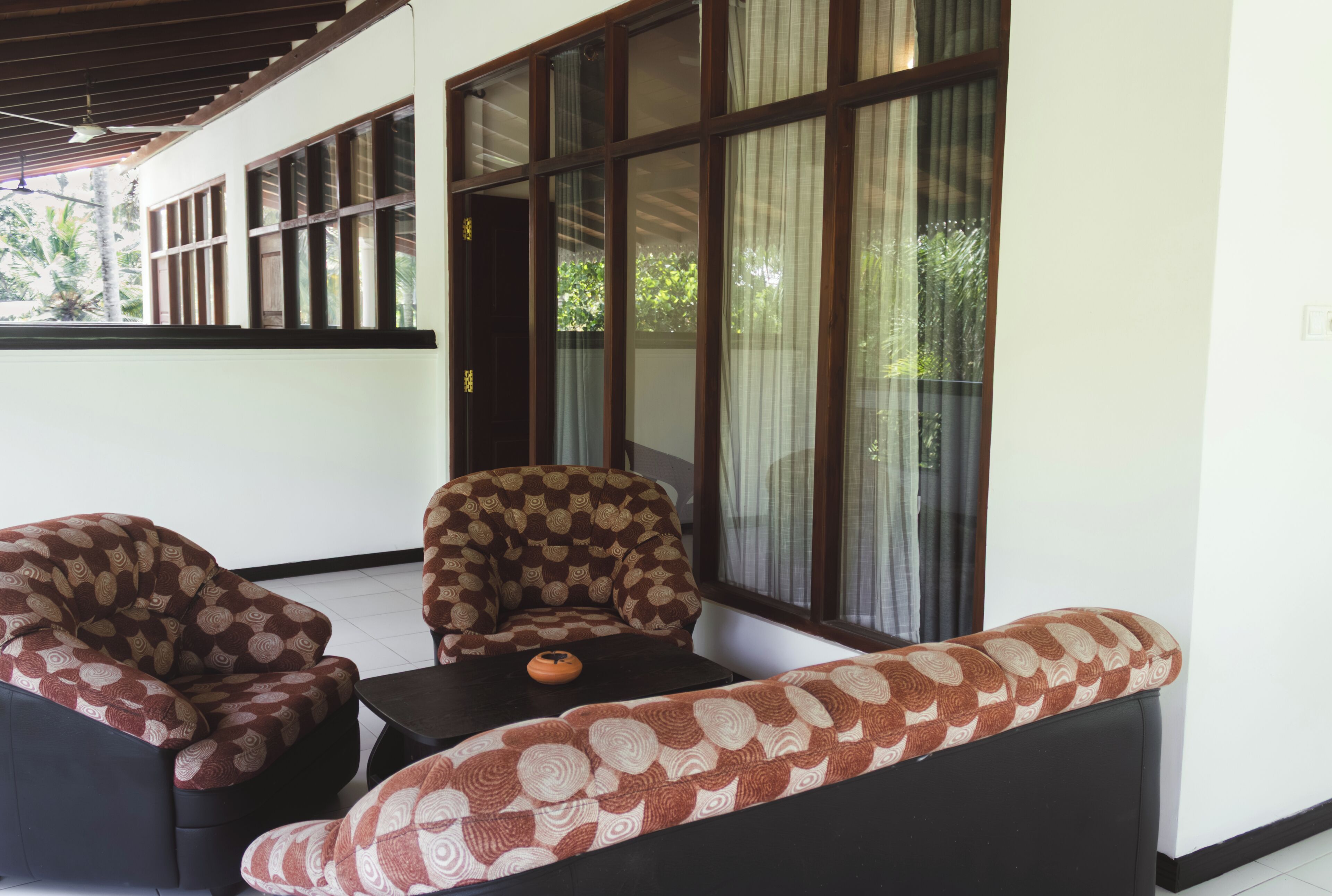 Una habitación con sofá, sillas, una mesita y ventanas amplias con cortinas.