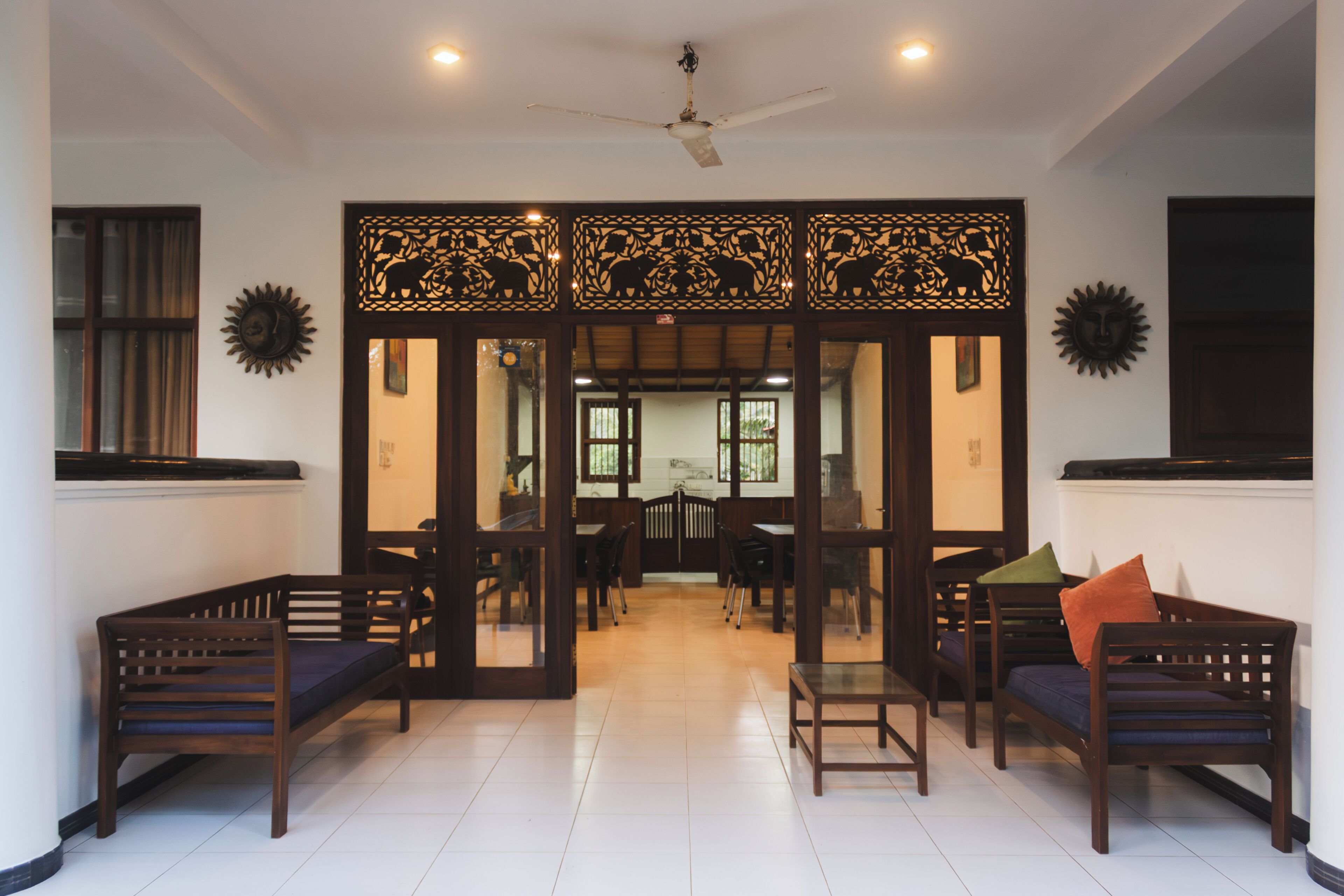 A room with a traditional design, featuring a wooden partition with animal carvings, a ceiling fan, and a seating area with wooden furniture.