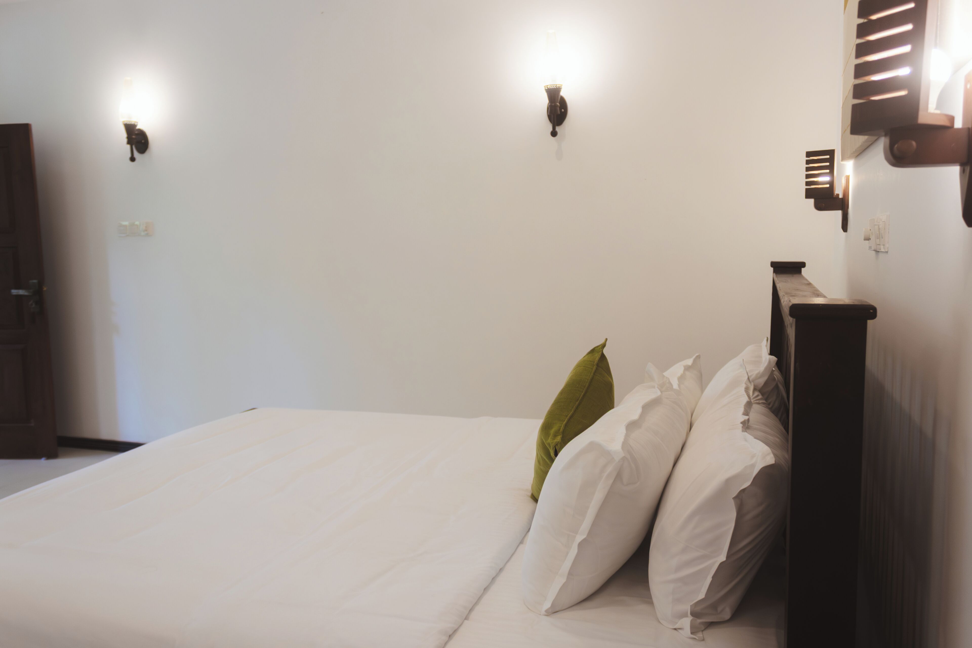 Una cama con ropa blanca y un cabecero de madera, dos lámparas de pared y una puerta a la izquierda.