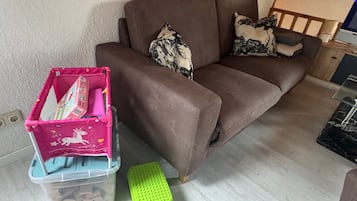 Ein braunes Sofa mit gemusterten Kissen, eine rosa Aufbewahrungsbox mit Einhorn-Motiv und eine grüne Trepfenbank.
