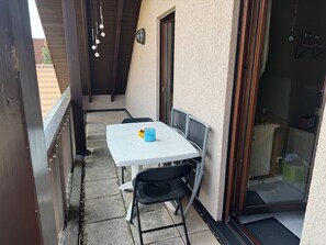 A balcony with a white table and two chairs, a small blue container, and a door leading to an interior space.