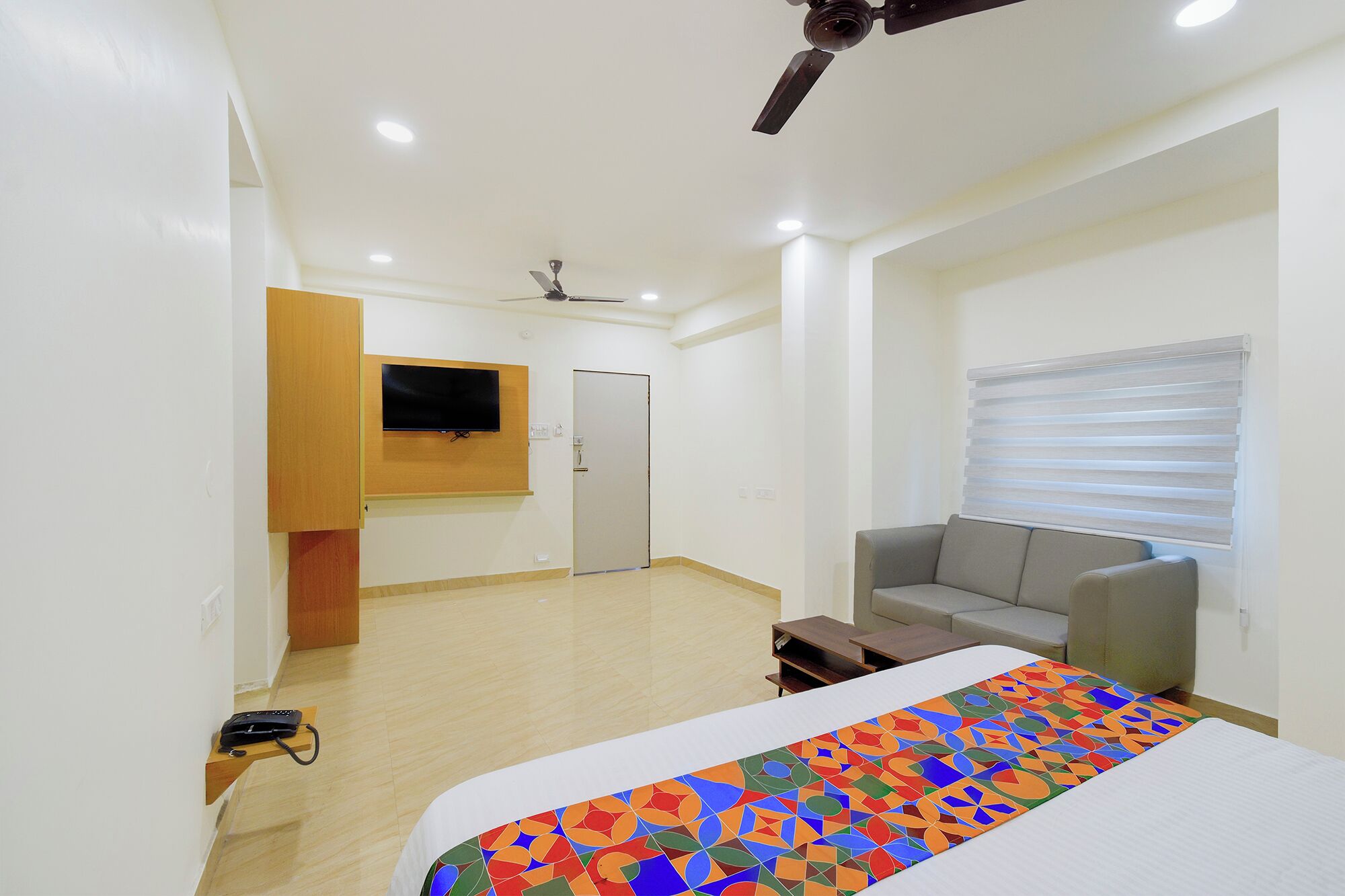 A hotel room with a bed, a sofa, a TV mounted on the wall, a ceiling fan, and a window with blinds.