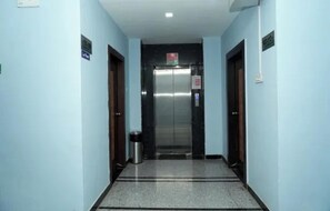 A hallway with a central elevator, light blue walls, and a tiled floor.