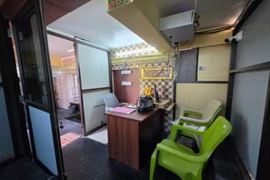 A small room with a desk, a chair, and a green plastic chair. There is a window with a view of a building outside. - Fabhotel Ar Residency Ii (Tiruvannamalai)