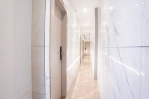 Corredor com paredes de mármore e chão de azulejos, que leva a uma porta com puxador.