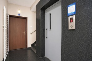Un pasillo con una puerta de madera, una puerta blanca con barandilla y un panel de control de ascensor fijado a la pared.