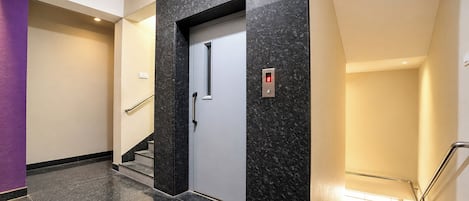 Un ascensor moderno con pantalla digital, pared de mármol y escalera con barandillas de metal.