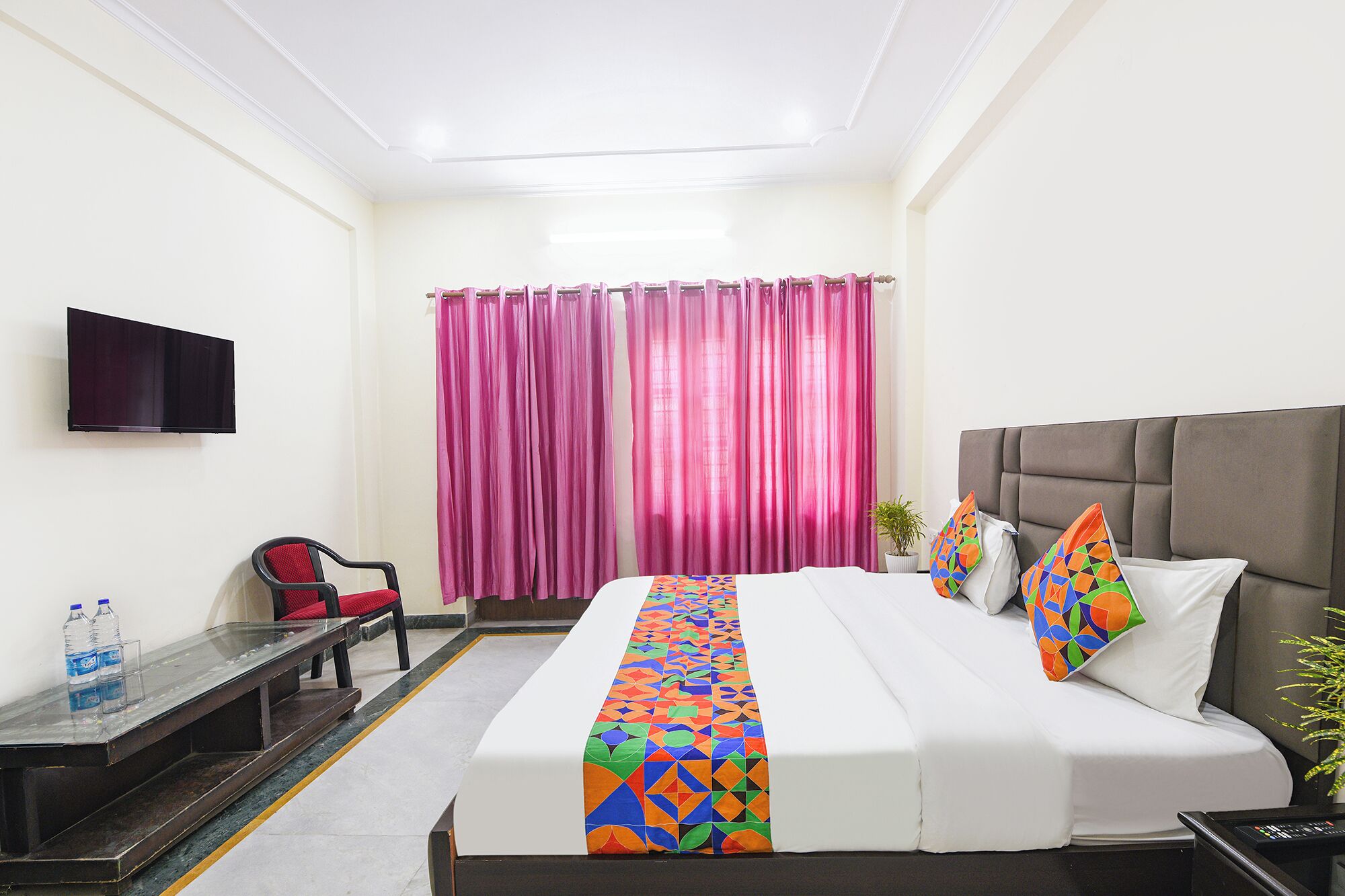 Habitación de hotel con cama, televisión montada en la pared, una silla, escritorio y ventana con cortinas rosadas.