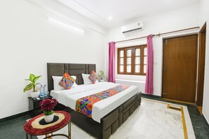 A hotel room with a bed, a bedside table with a vase of flowers, a window with curtains, and a wooden door. - Fabhotel Maurya Residency (Gurugram)