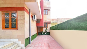 Un sendero estrecho que conduce a un edificio con una puerta roja y una ventana. Hay escalones que suben a la entrada, y un seto verde a la derecha.
