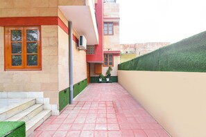 Un sendero estrecho que conduce a un edificio con una puerta roja y una ventana. Hay escalones que suben a la entrada, y un seto verde a la derecha.