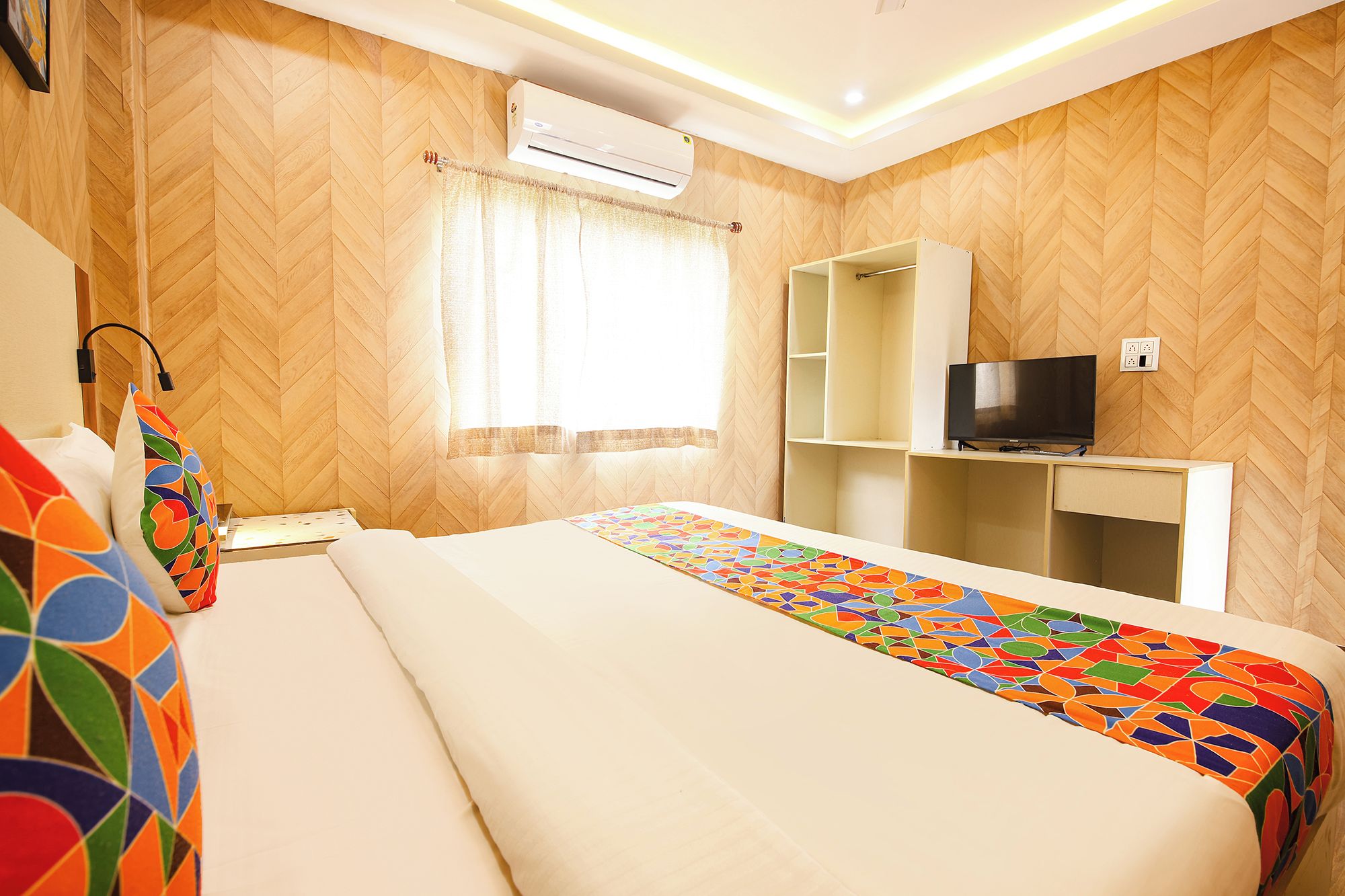 Habitación de hotel con una cama, un televisor sobre un mueble, una estantería y un equipo de aire acondicionado.
