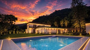 Una piscina con borde de azulejos, rodeada de un área exterior bien iluminada con una casa y árboles.