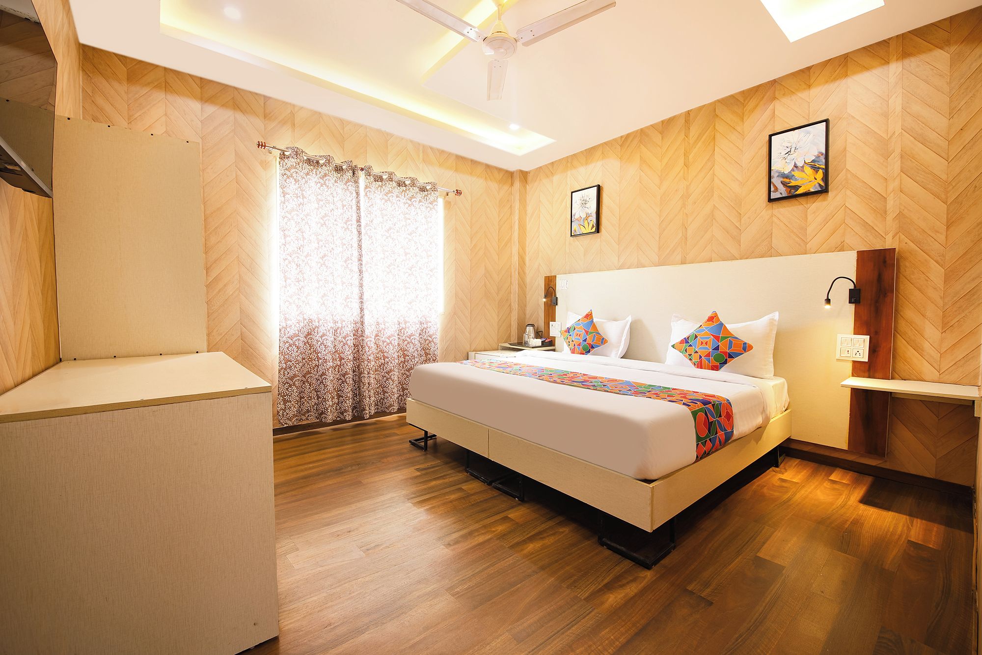 Habitación de hotel con cama, mesita de noche, lámpara de pared, ventilador de techo y ventana con cortinas.