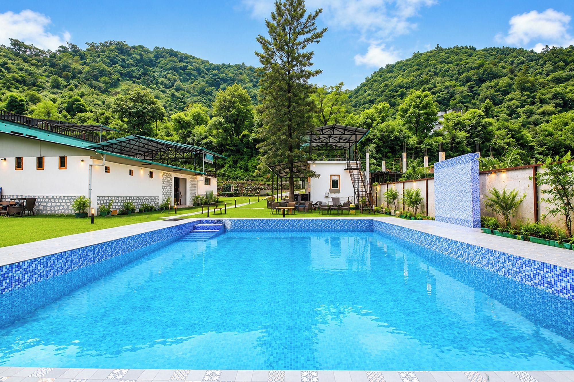 Una piscina con borde de azulejos, rodeada por un edificio blanco con techo verde, y un paisaje montañoso boscoso al fondo.