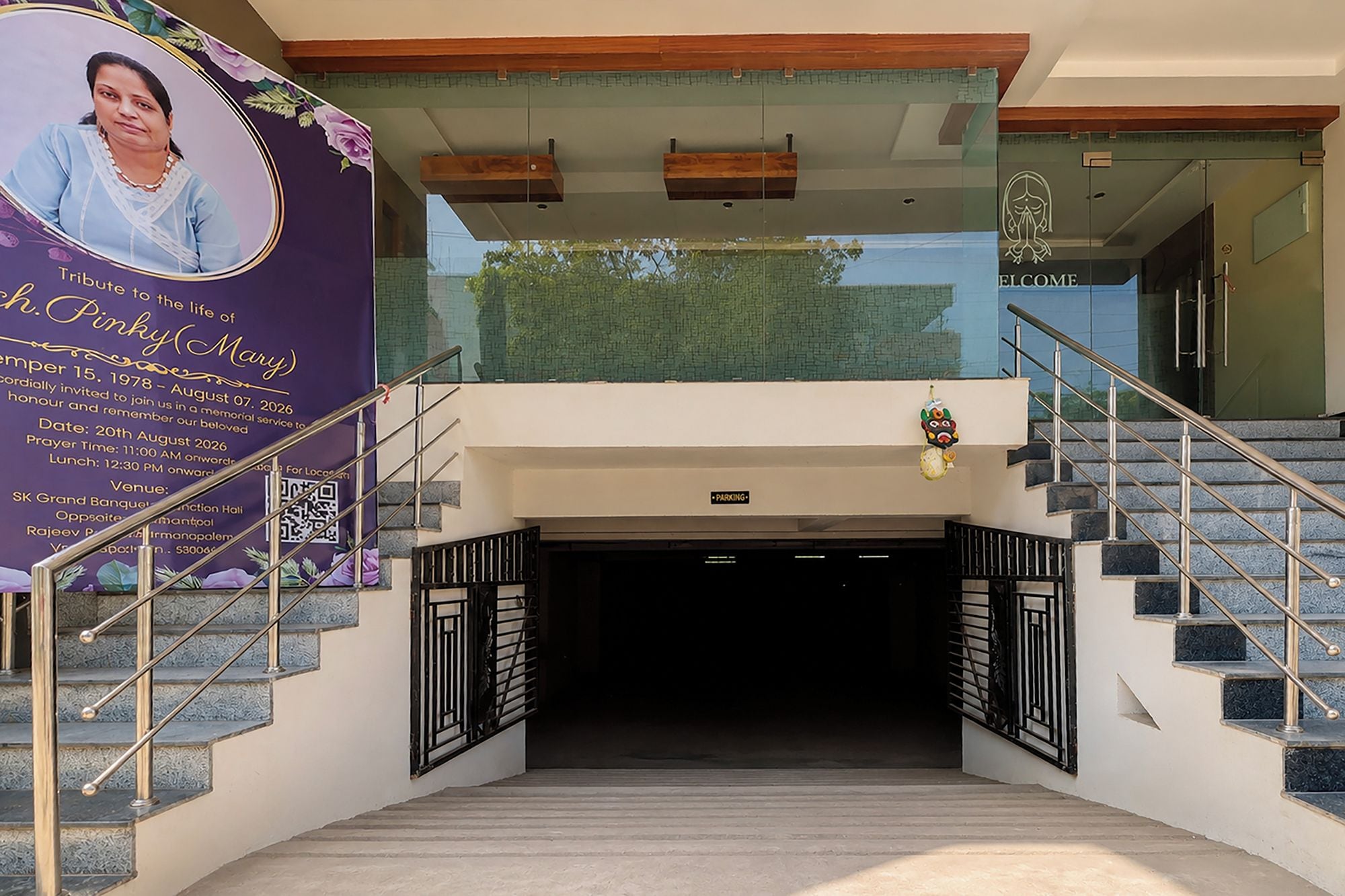 Entrada de hotel con fachada de vidrio, una escalera con barandales metálicos y un cartel de bienvenida.