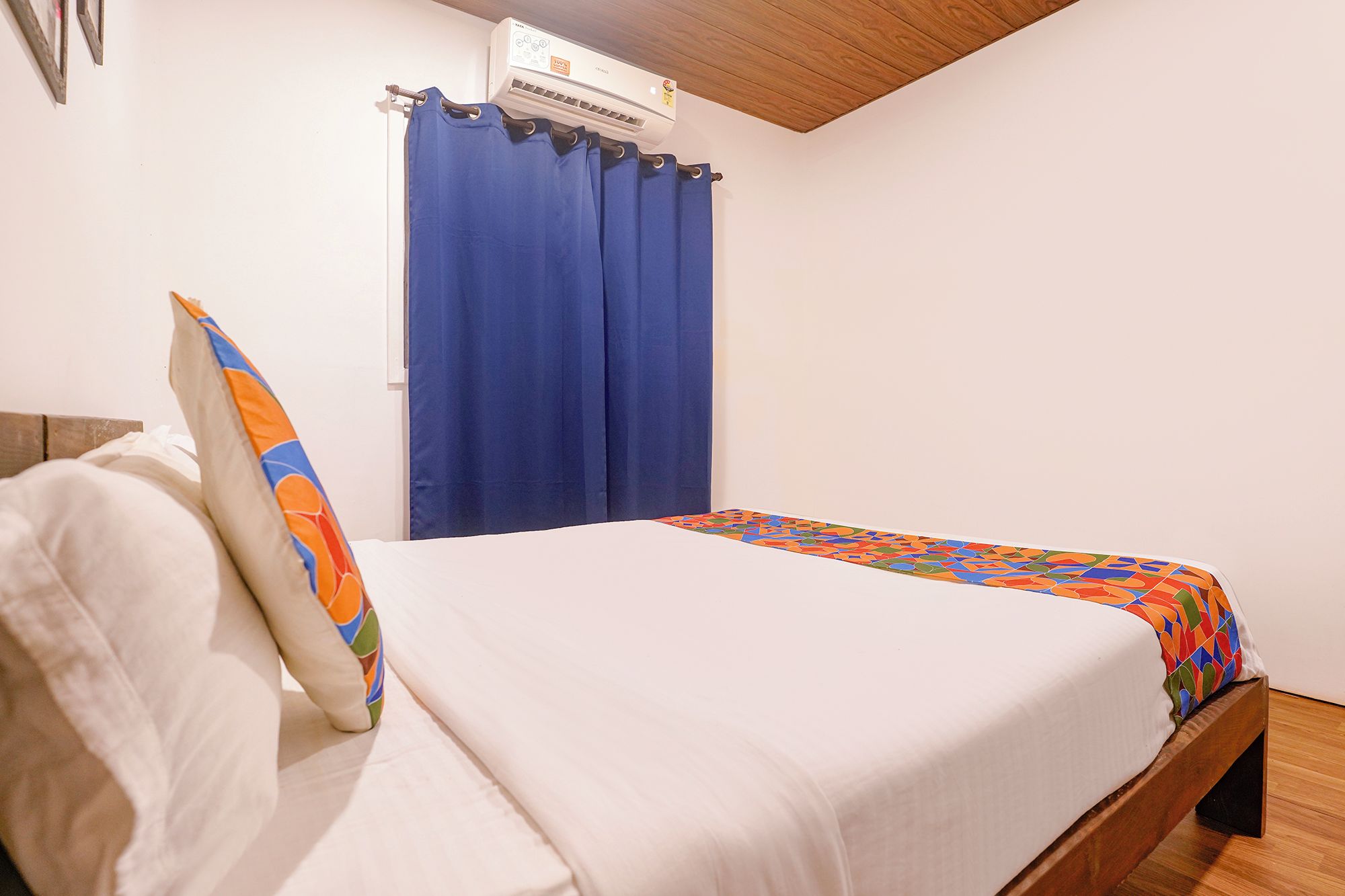 Una cama individual con colchón blanco y ropa de cama de colores, una cortina azul y un ventilador de techo.