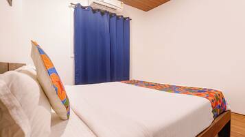 Una cama individual con colchón blanco y ropa de cama de colores, una cortina azul y un ventilador de techo.