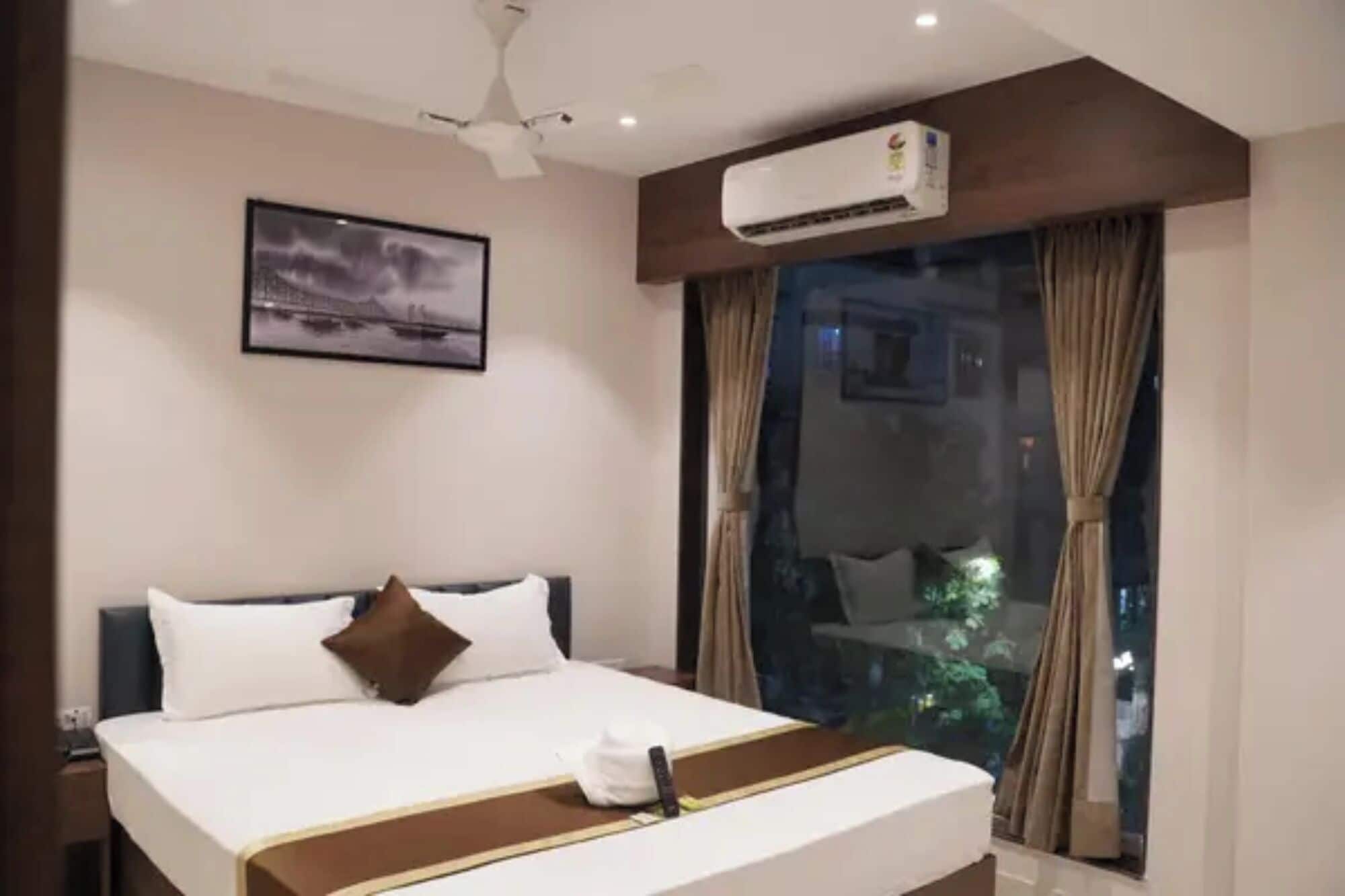 A hotel room with a bed, a ceiling fan, an air conditioning unit, a framed picture on the wall, and a window with curtains.