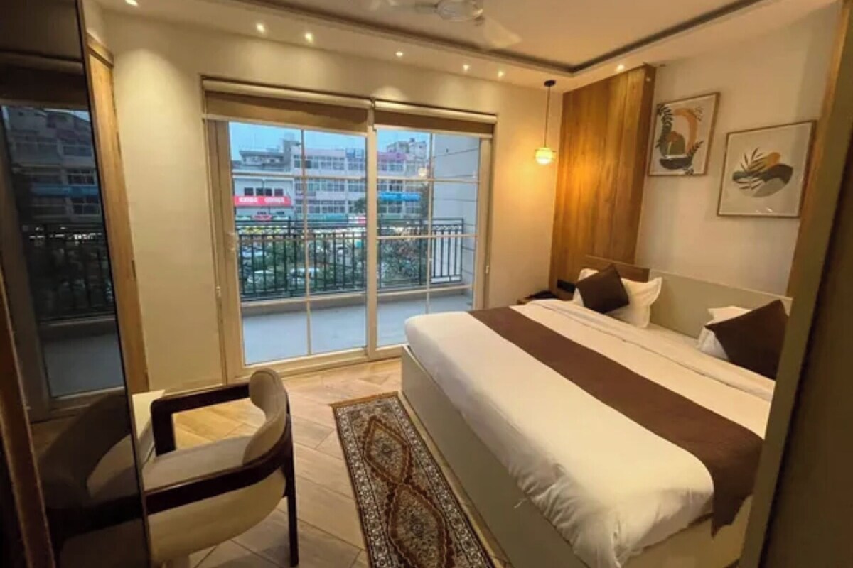 Un dormitorio con una cama grande, una silla, un balcón con vistas a edificios y un ventilador de techo.