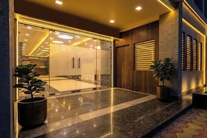 Entrada de un hotel con puerta de cristal, plantas en macetas y piso de baldosas.
