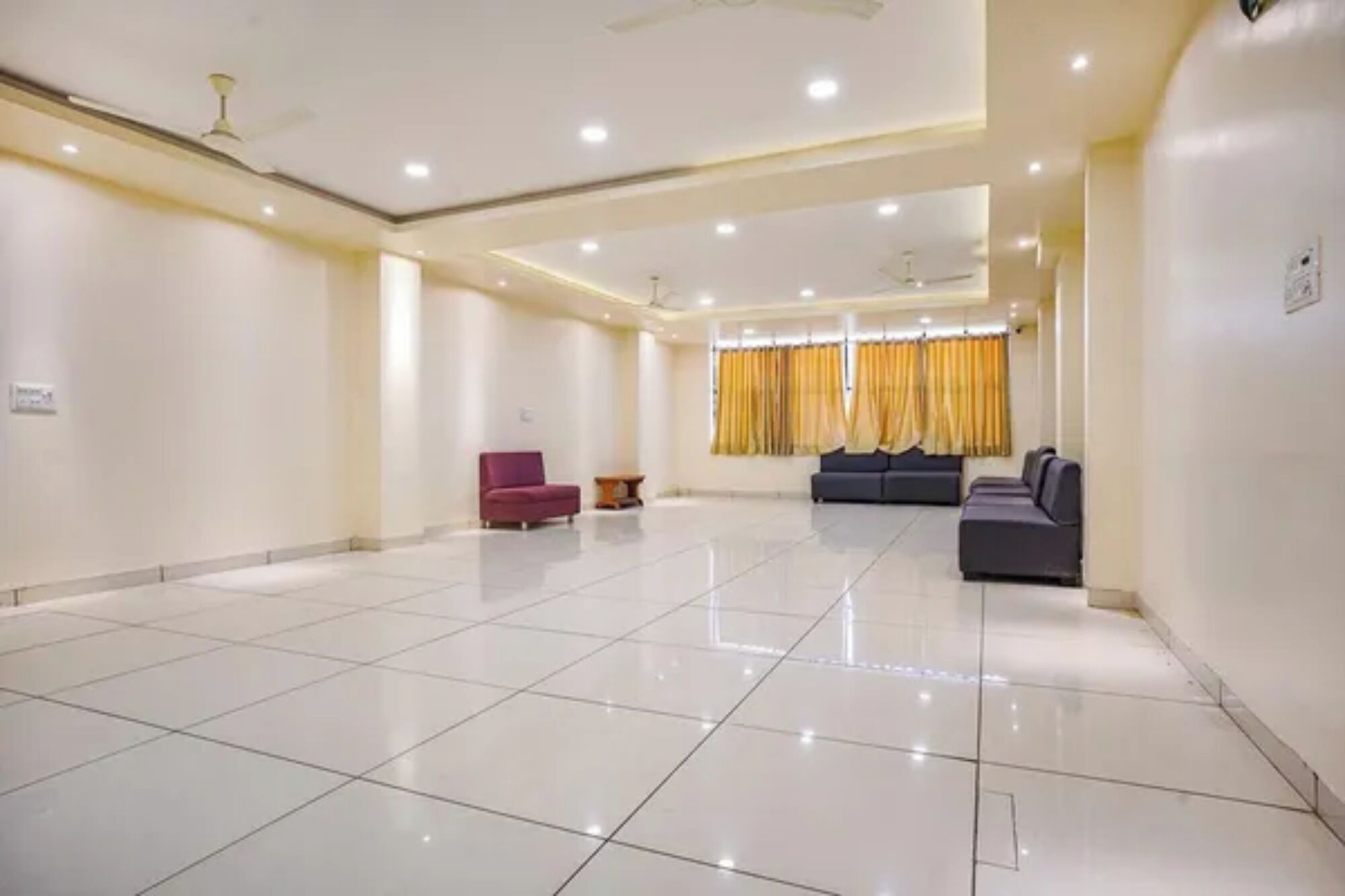A spacious, well-lit room with a tiled floor, a red chair, a dark sofa, and a window with yellow curtains.