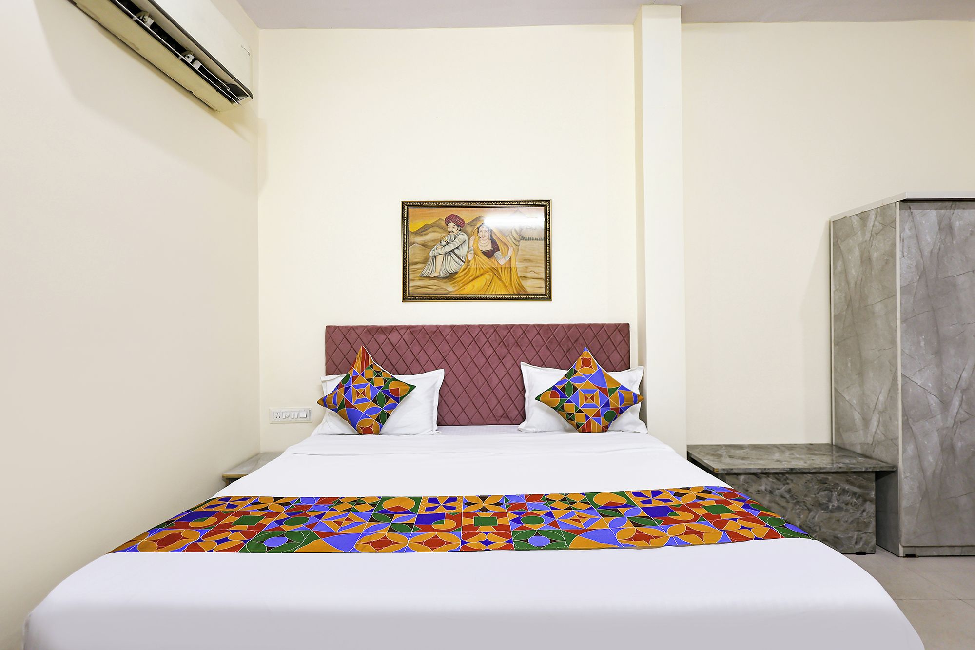 Una habitación de hotel con una cama, dos almohadas, un cuadro colgado en la pared, una mesita de noche de mármol y un banco de mármol.