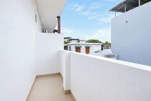 Un pasillo estrecho y blanco con piso de baldosas que conduce a un balcón con vista a un área residencial con edificios tradicionales.