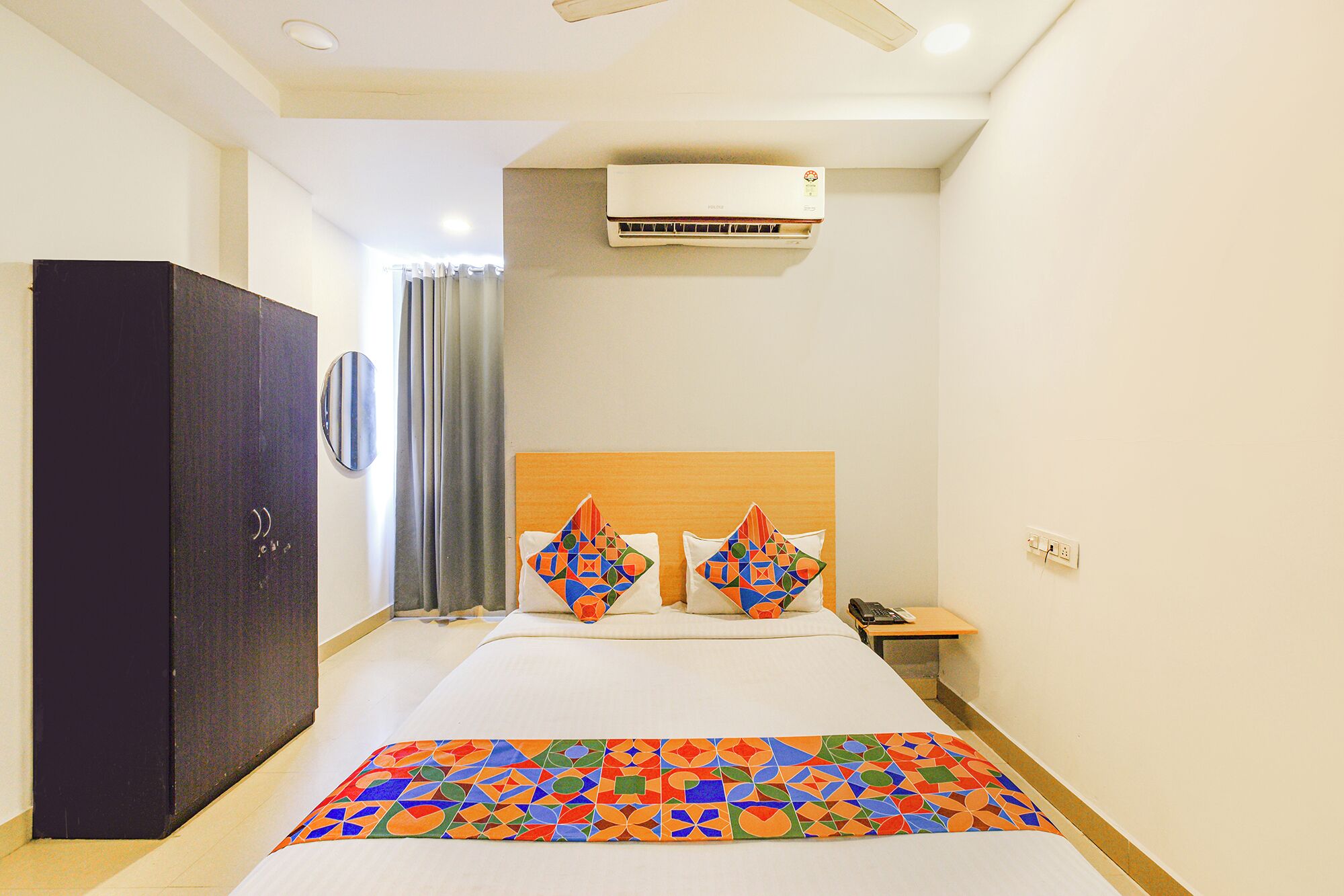 A hotel room with a bed, a wardrobe, an air conditioning unit, a bedside table with a phone, and a wall-mounted mirror.