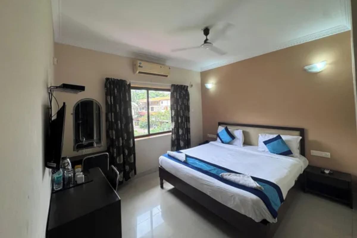 A hotel room with a bed, a desk, a chair, a window with curtains, a ceiling fan, and an air conditioning unit.