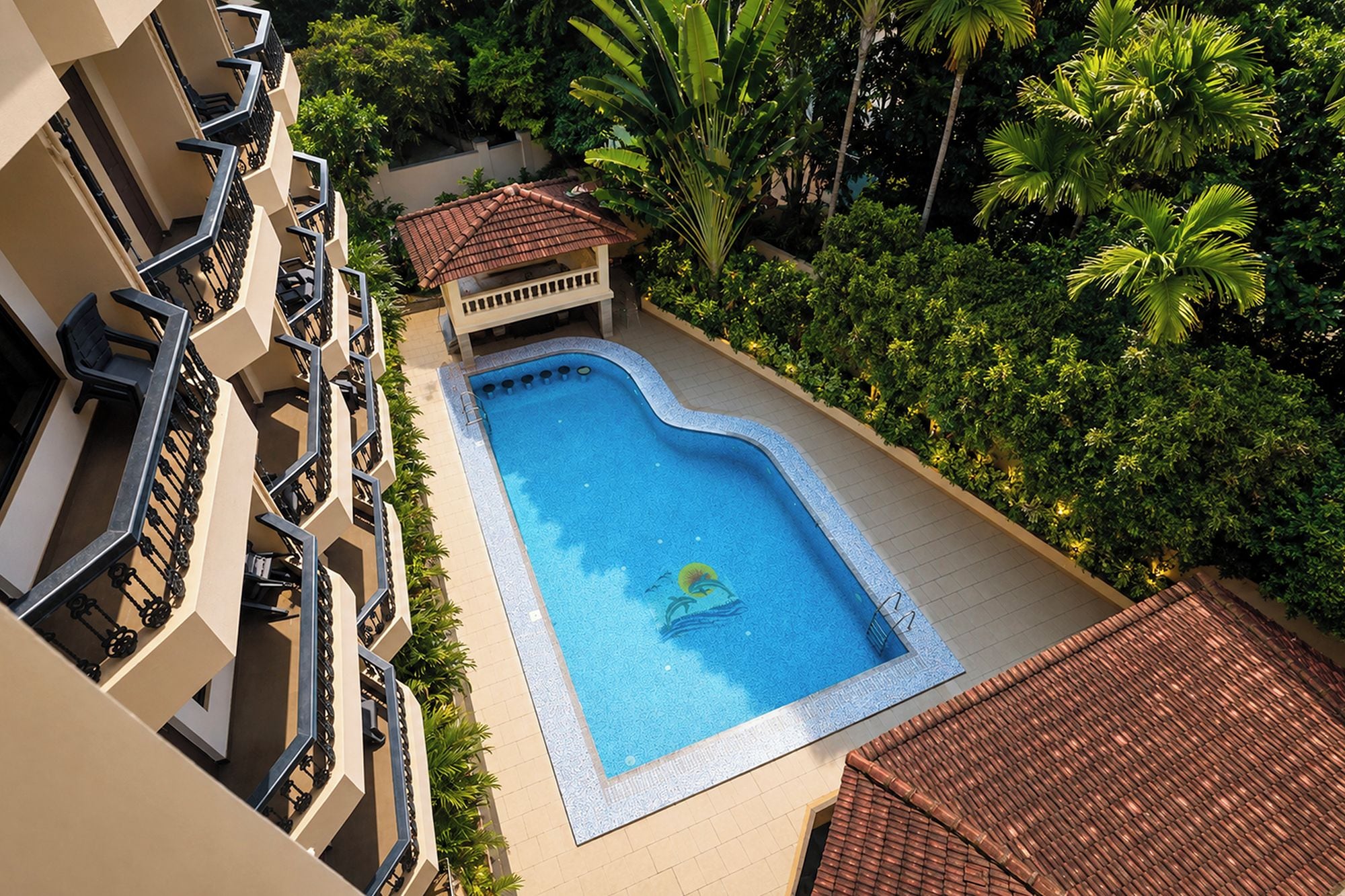 A swimming pool with a tiled deck, surrounded by lush greenery and a building with balconies.
