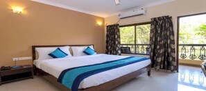 A hotel room with a bed, a nightstand, a window with curtains, and an air conditioning unit. - Oriva Rendezvous Beach Resort (Panaji)