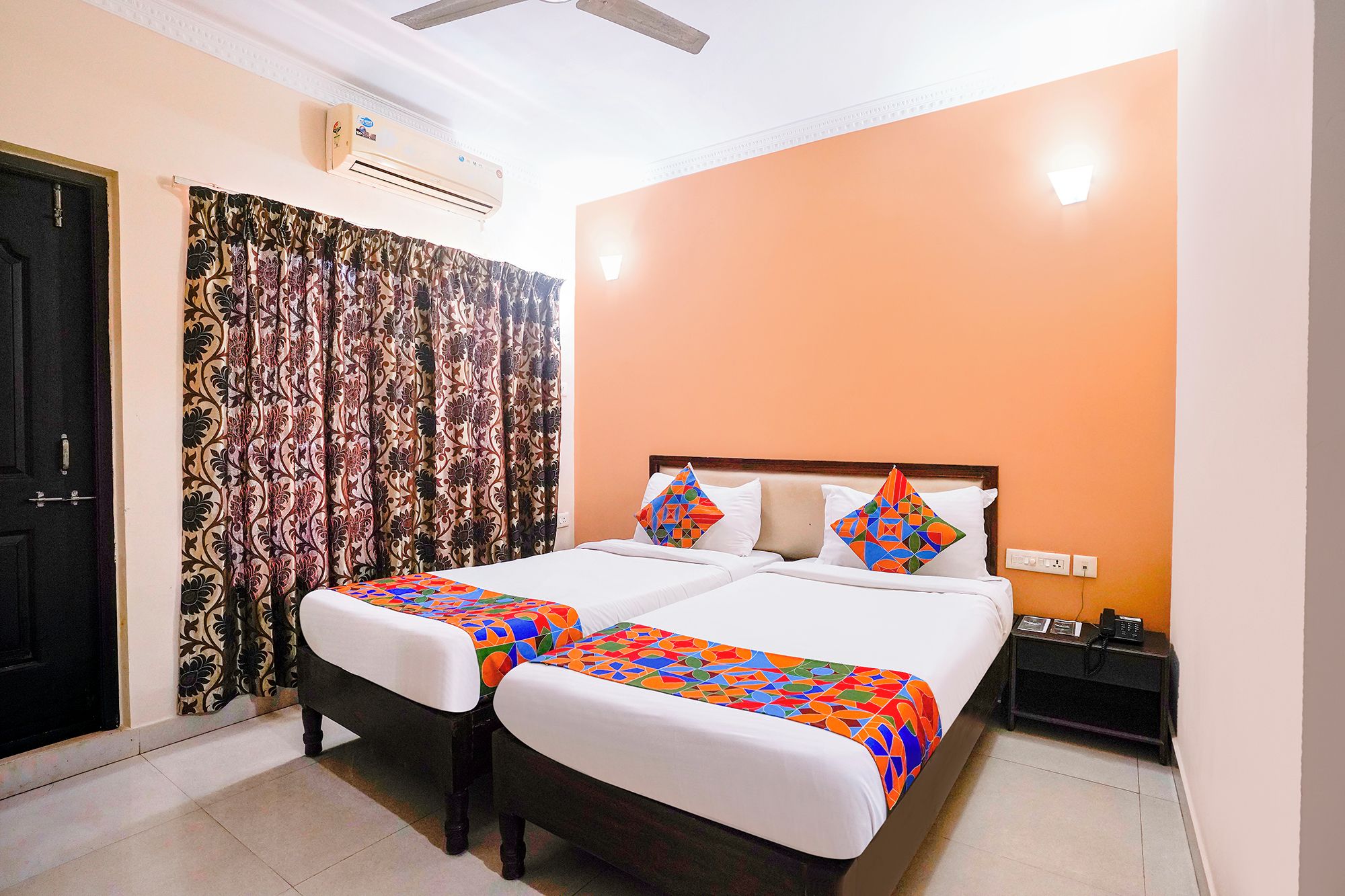A hotel room with a bed, a bedside table, an air conditioning unit, and a door.