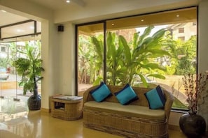 A wicker sofa with cushions, a small side table, and a large window with a view of greenery. - Oriva Rendezvous Beach Resort (Panaji)