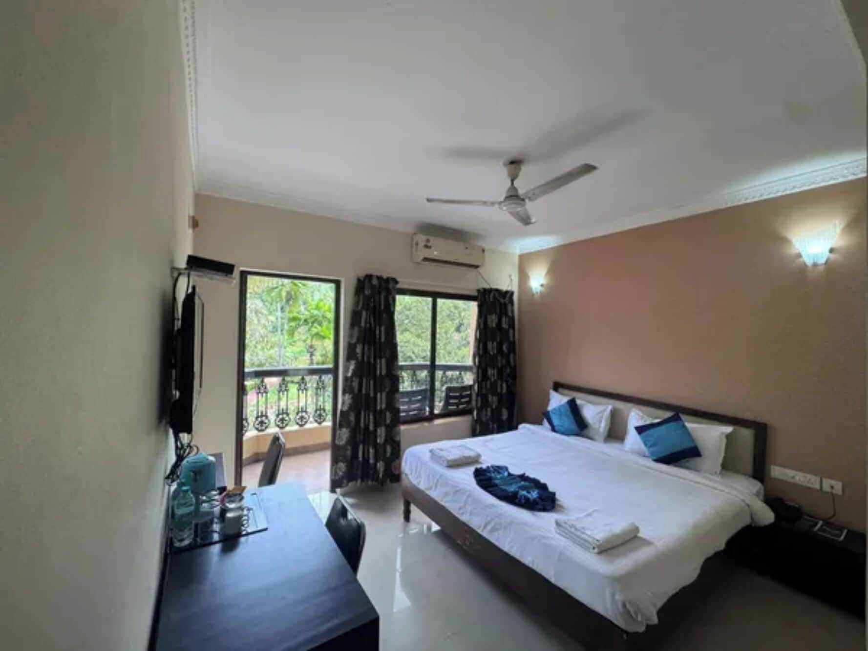 Una habitación de hotel con cama, escritorio, silla, ventilador de techo y balcón con cortinas.
