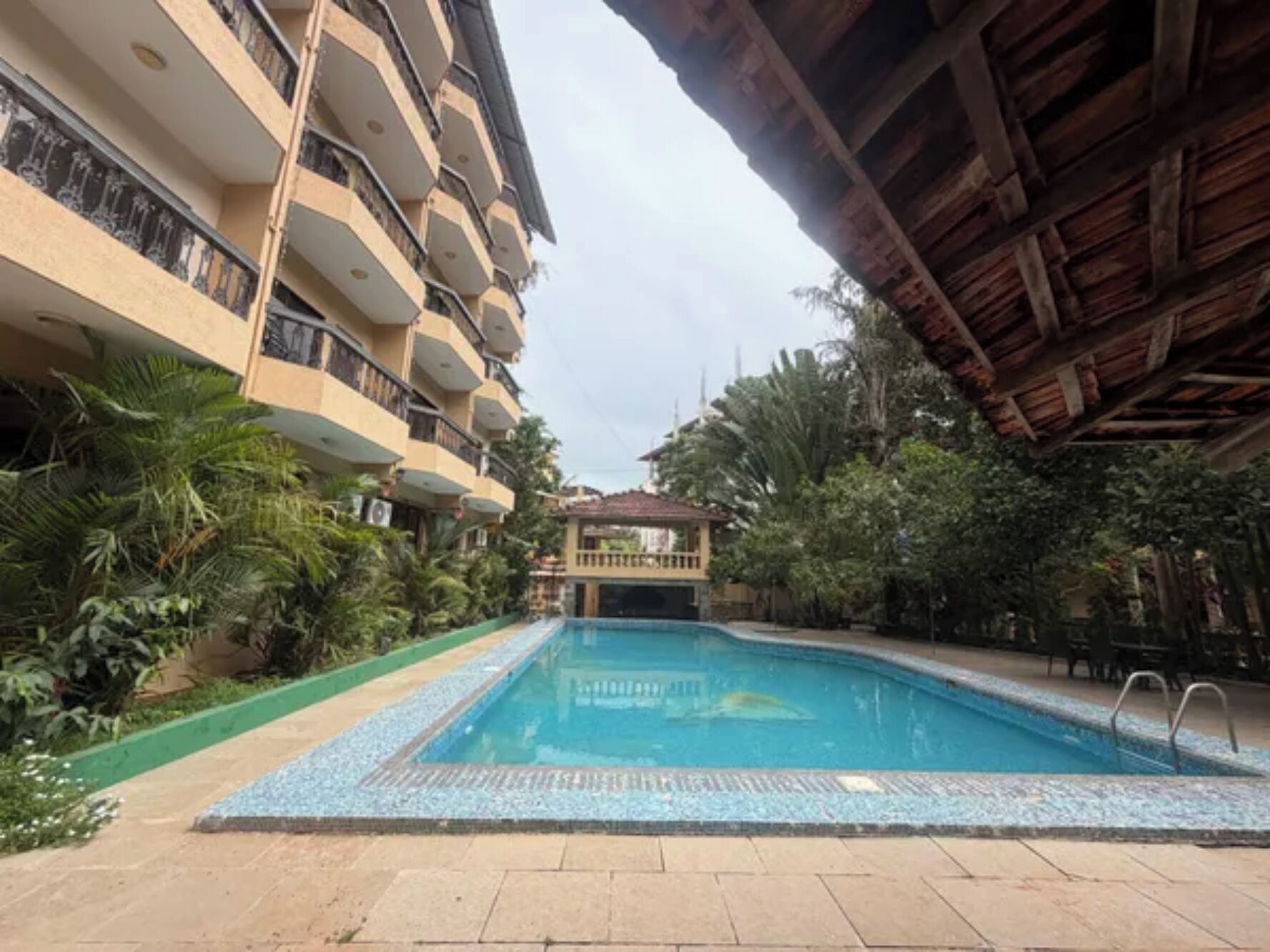 Una piscina con borde de azulejos, rodeada de vegetación y un edificio de varios pisos con balcones.