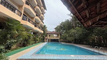 Una piscina con borde de azulejos, rodeada de vegetación y un edificio de varios pisos con balcones.