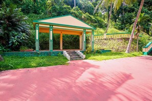 A covered pavilion with a green and beige color scheme, surrounded by lush greenery and a red pathway leading to it.