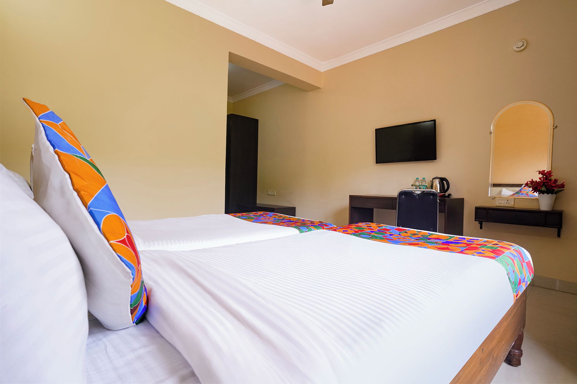 Quarto de hotel com duas camas, televisão, secretária com espelho e planta em vaso.