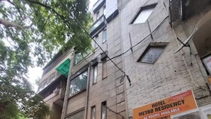 Edifício com placa indicando 'Hotel Metro Residency' e quartos disponíveis. Uma motocicleta está estacionada na frente e um vaso de planta próximo à entrada.