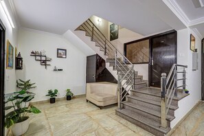 A spacious living area with a beige sofa, a staircase with a glass railing, and a door marked with the number 002. - FabHotel Galleria Stayz (Gurugram)