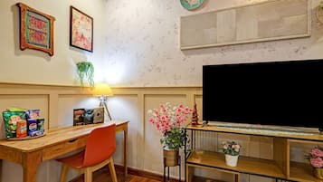Huoneessa on puinen työpöytä, punainen tuoli, jalustalla oleva televisio sekä koriste-esineitä, kuten kehystettyjä valokuvia ja seinälle kiinnitetty kasvi.
