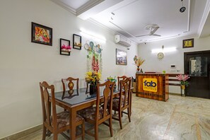 A hotel reception area with a reception desk, a dining area with a glass-top table and wooden chairs, and wall-mounted decorations including framed artwork and a clock. - FabHotel Galleria Stayz (Gurugram)